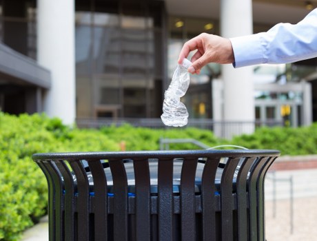 A person's hand is shown dropping a clear plastic bottle into a black metal rubbish bin located outdoors. The bin is positioned on a paved surface with some green shrubbery visible in the background, alongside a building with glass windows and structural elements suggesting an urban or commercial environment. The hand is wearing a light blue long-sleeved shirt, and the plastic bottle appears to be empty, with a slight reflection on its surface. The scene reflects waste disposal activity, consistent with rubbish removal services in the East Ham area, emphasizing proper disposal of plastic waste in a typical street or public space setting.