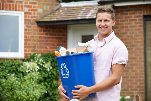 A young man with light brown hair, wearing a light pink short-sleeved shirt, stands outdoors in front of a brick house with a white window and tiled roof. He is smiling and holding a blue recycling bin filled with various types of rubbish, including cardboard boxes, paper, plastic bottles, and miscellaneous waste. The bin has a white recycling symbol on the side. In the background, there are green bushes with white flowers, and the scene appears to be during daytime with natural lighting, indicating a tidy garden or driveway area in East Ham, suitable for rubbish collection or garden clearance services from Garden Clearance East Ham.