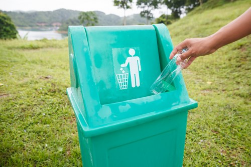 A hand placing a clear plastic bottle into a bright turquoise recycling bin with a white icon of a person depositing waste, situated outdoors on a grassy area with a backdrop of trees and distant water, indicating an organized waste collection effort typical of environmental initiatives in East Ham. The bin's smooth, glossy plastic surface contrasts with the textured green grass on the ground, and natural daylight illuminates the scene, emphasizing the cleanliness and civic responsibility associated with rubbish removal and recycling services offered by Garden Clearance East Ham in the local area.