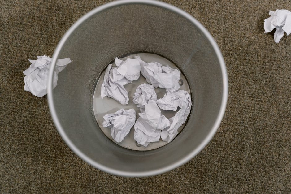 A cylindrical metal rubbish bin with a smooth, matte silver finish sits on a brown, textured carpeted floor. Inside the bin, several crumpled sheets of white paper are visible, partially submerged in 