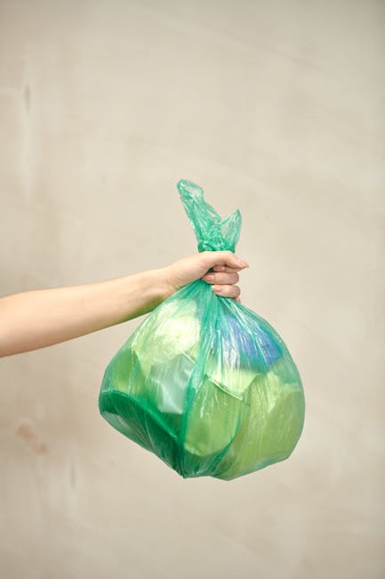 A person's hand and forearm are visible, holding a green plastic rubbish bag by its twisted knot at the top. The bag appears to contain various waste materials, with some blue and yellow hues visible