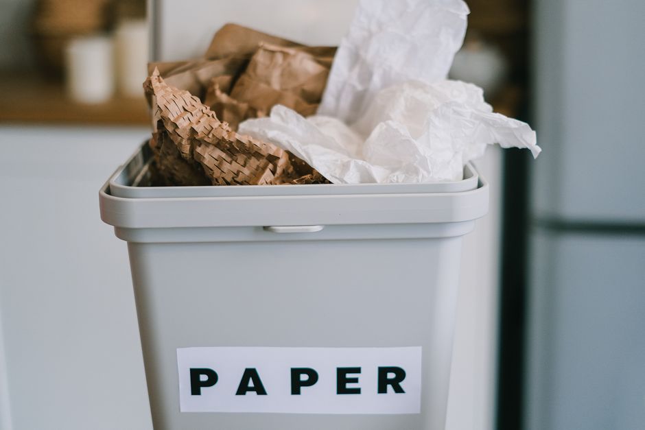A pile of mixed household waste including flattened cardboard boxes, crumpled paper, and plastic packaging is gathered at the base of a brick and concrete wall, partially sheltered by a large tree tru