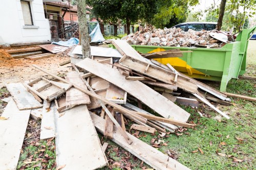 A pile of discarded wooden floorboards and planks, varying in size and shape, with some appearing weathered and aged, stacked loosely in front of a bright green skip filled with mixed rubble and debris on a grassy outdoor area. In the background, there are trees, a paved pathway, and a residential building with a small staircase, indicating a garden or backyard setting, possibly in East Ham. The scene suggests ongoing garden clearance or renovation work, with visible signs of construction or demolition waste. Garden Clearance East Ham provides rubbish removal services suitable for clearing such waste from residential or outdoor spaces in the area.