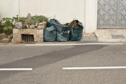 Four large green refuse sacks filled with garden waste and household rubbish are stacked on the pavement next to a low brick wall with some small plants and rocks in front of it. The sacks are slightly crumpled and tied at the top, with some stems and debris protruding from one of them. The rubbish is positioned alongside a cream-colored wall and a decorative metal gate on the right. The street in front has a dark asphalt surface marked with white lines, indicating a parking or traffic lane, and the scene appears to be in a residential area within or near East Ham. The setting suggests a typical garden clearance or rubbish collection context, relevant to professional rubbish removal services offered by Garden Clearance East Ham.