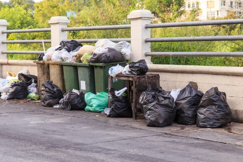A collection of black and green rubbish bags filled with household waste stacked along a paved urban sidewalk, with some bags leaning against a row of waste bins and others placed directly on the ground. The waste includes plastic and paper materials visible through the loosely closed bags. Behind the bags, there is a metal railing and a concrete bridge with greenery and trees in the background, indicating an outdoor setting possibly near a park or residential area in East Ham. The scene suggests ongoing rubbish collection or disposal activity, relevant to garden clearance and waste removal services offered by Garden Clearance East Ham in the local postcode area.